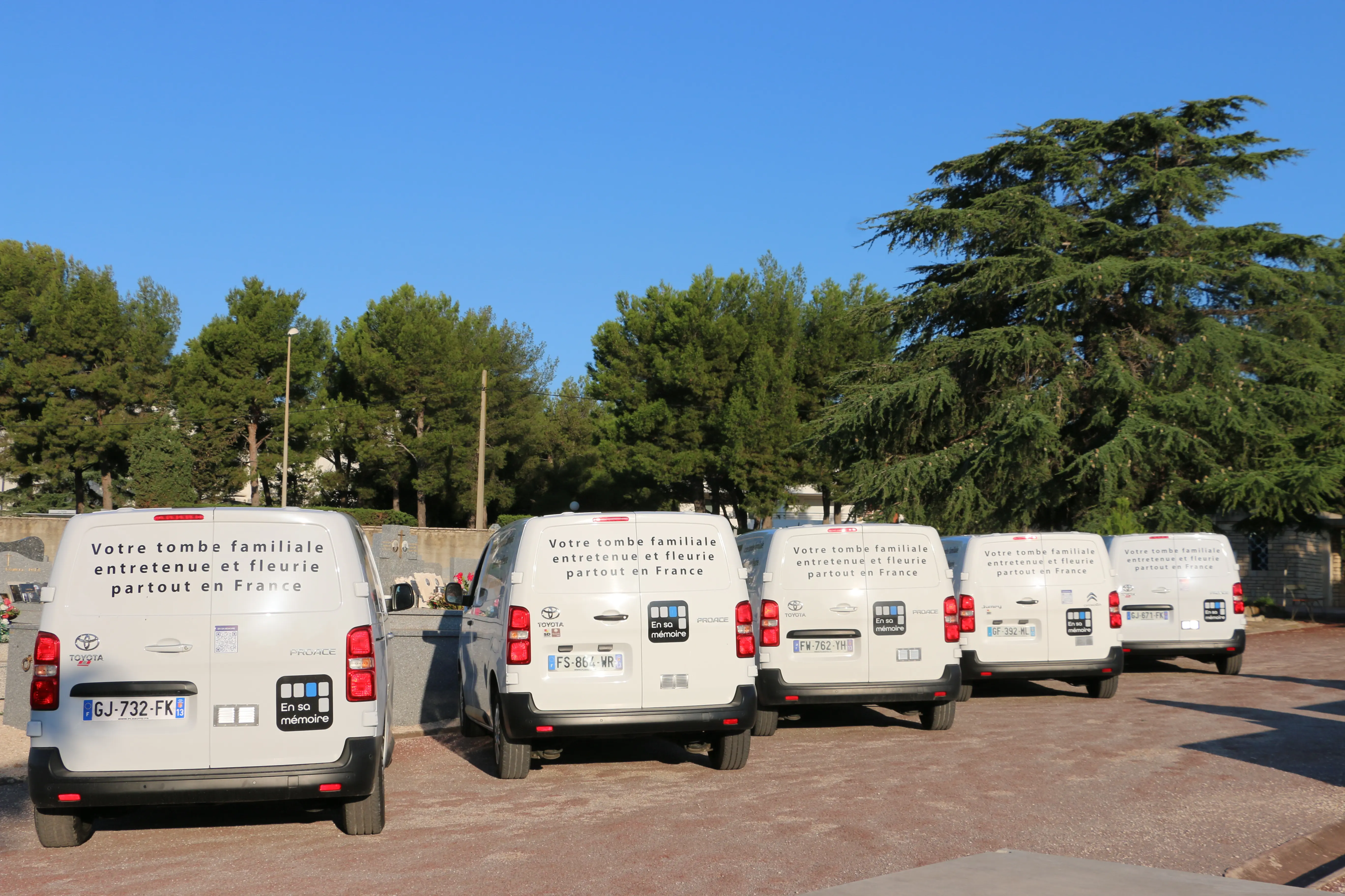 Vans blanches alignées dans un parking près d’arbres, messages indiquant une tombe familiale entretenue et fleuri en France.