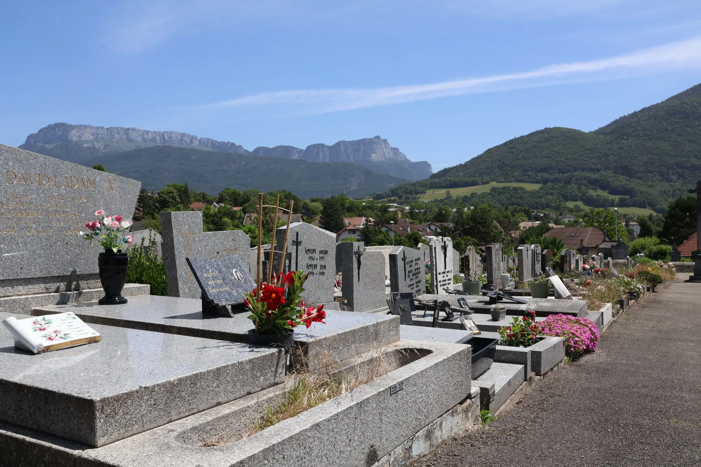 Allée de tombes en pierre au cimetière, fleurs et monuments gravés, montagnes en arrière-plan sous ciel clair. intégrant le sujet : comment nettoyer la tombe d'un proche.