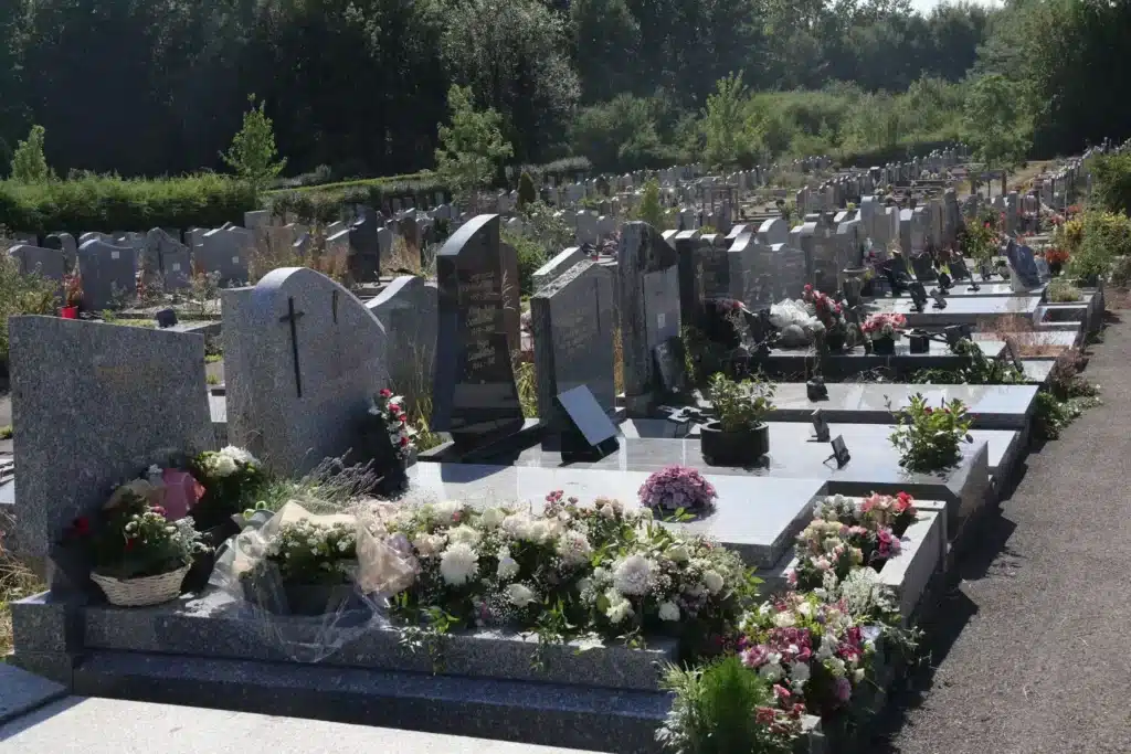 Ligne de tombes gravées et monuments funéraires dans un cimetière communal, entourés de fleurs et de sentiers, avec verdure au loin. Ce qui est obligatoire dans un cimetière fréquente les clôtures et ossuaire.