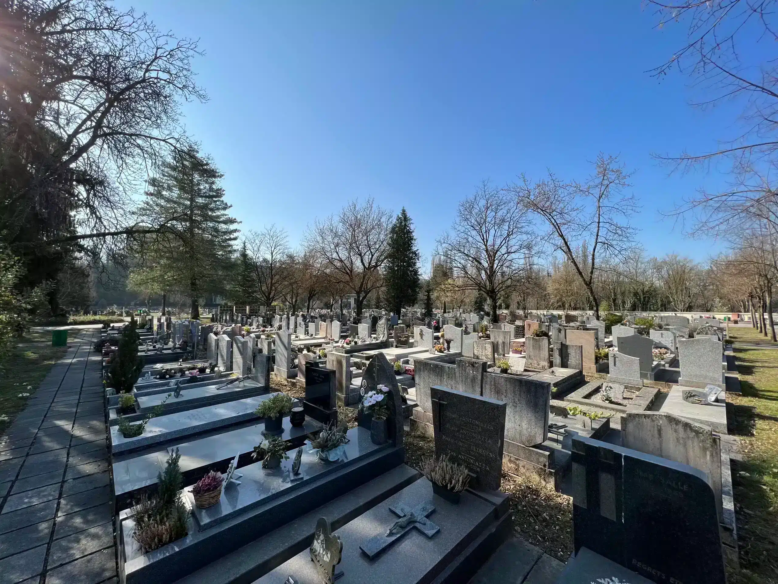 Cimetière lumineux avec de nombreuses tombes en rangées, pierres tombales et croix; arbres nus et chemin pavé à gauche dans un ciel bleu. Acte notarié de donation concession funéraire évoqué.