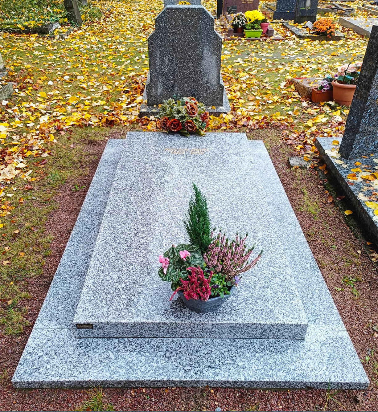 Tombe en pierre grise ornée de fleurs artificielles et de plantes, posé dans un cimetière avec des feuilles jaunes au sol.