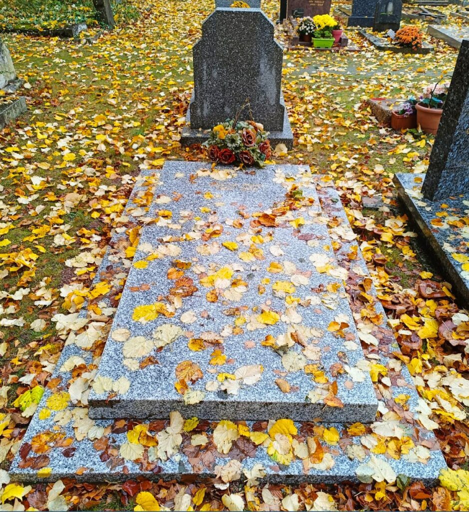 Tombe au cimetière recouverte de feuilles d’automne, avec bouquet posé près de la pierre.