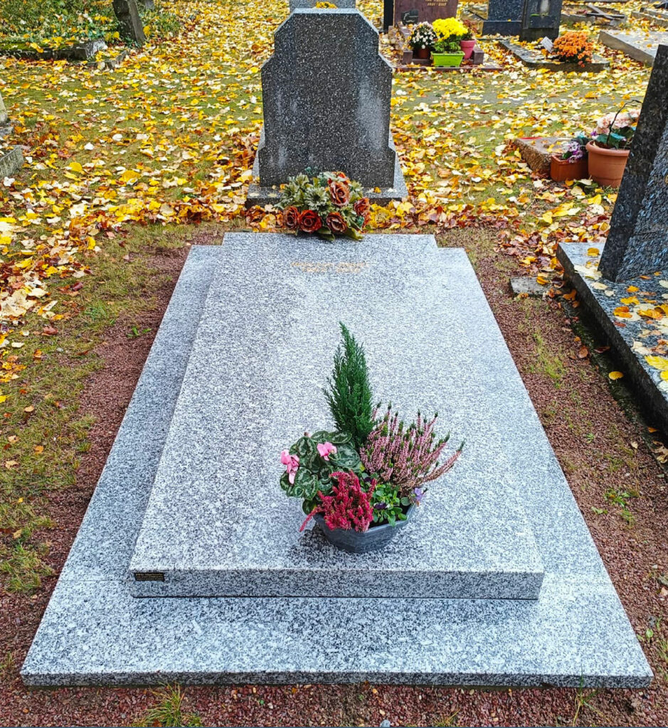 Tombe en pierre grise avec une urne florale sur le socle, entourée de feuilles tombées et d’autres monuments funéraires en
